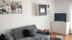 A modern living room with a grey sofa, a coffee table, a TV on a white cabinet, and a painting of flowers on the wall.