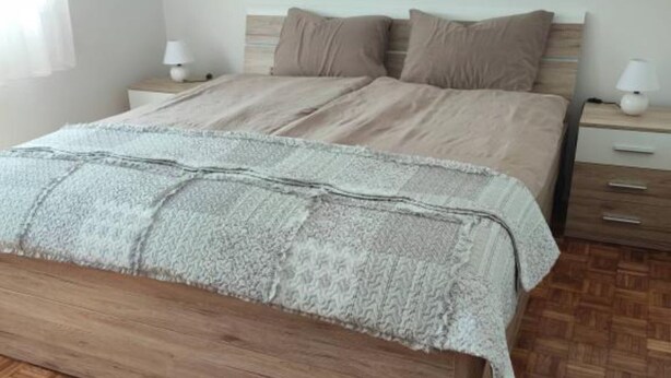 A bedroom with a wooden bed, a white blanket, two bedside tables with lamps, and a herringbone-patterned floor.