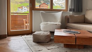 Ein Wohnzimmer mit Holzboden, einem beigefarbenen Sofa, einem weißen Sessel, einem Holztisch und Blick auf eine Berglandschaft durch große Fenster.