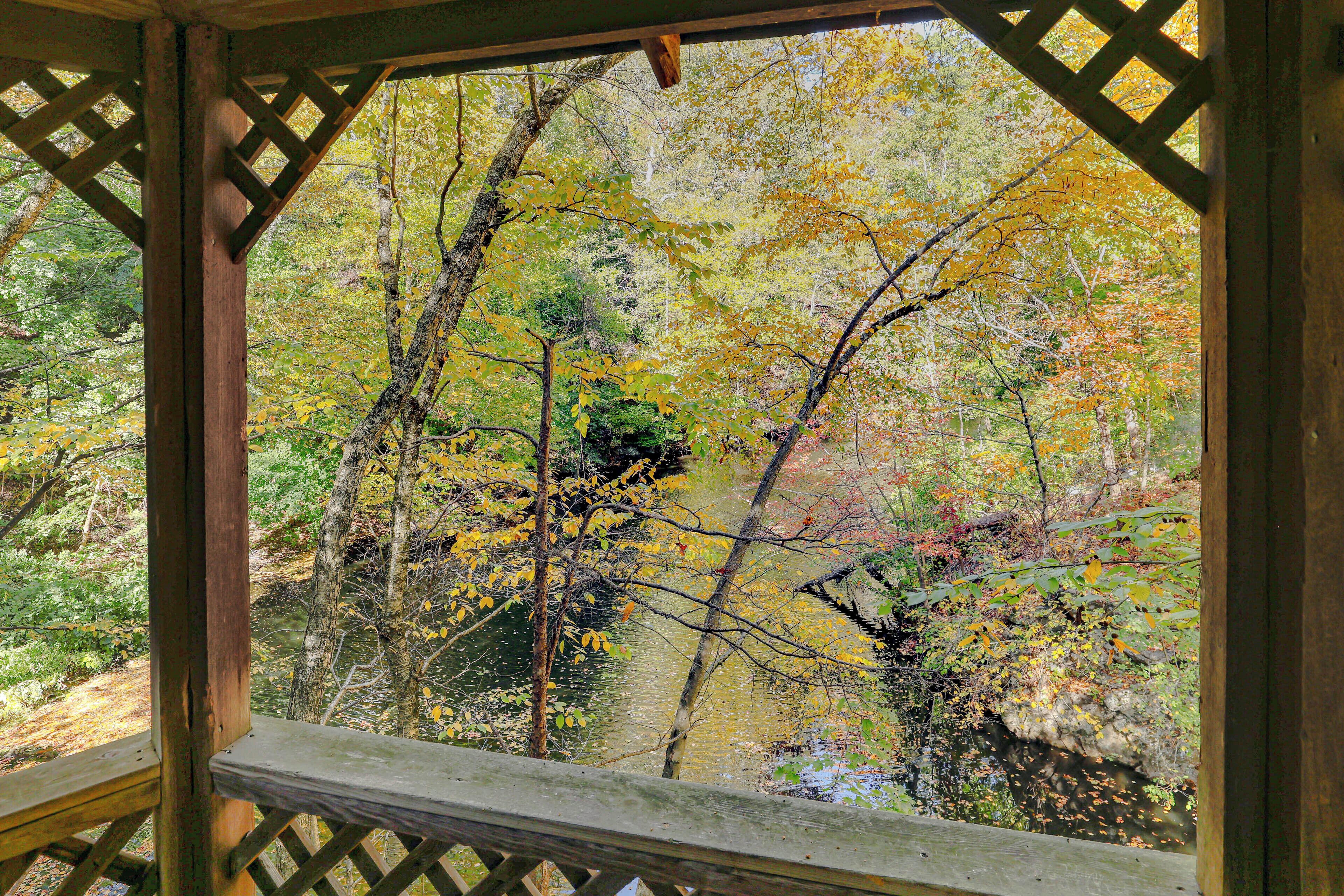 A wooden gazebo with a lattice roof overlooking a forested area with a stream.