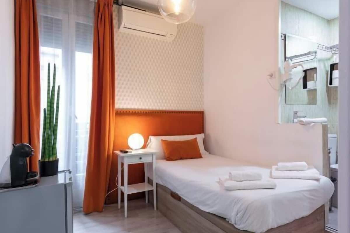 A hotel room with a bed, a bedside table with a lamp, a potted plant, and a bathroom visible through an open door.