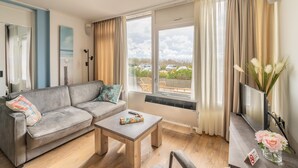 A modern living room with a grey sofa, a wooden coffee table, and a large window with curtains. - 2 Person Island View Apartment (De Koog)