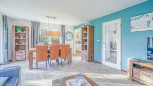 A spacious living room with a dining area, wooden furniture, a large clock, and a TV on the right. - Charming Villa Near the sea and Forest (De Koog)