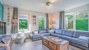 A modern living room with a sectional sofa, a wooden coffee table, and large windows offering a view of greenery. - Charming Villa Near the sea and Forest (De Koog)