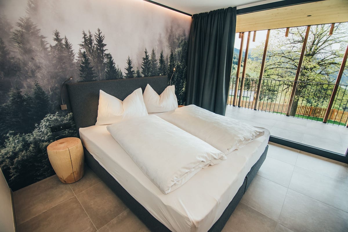 A modern bedroom with a large bed, a view of trees through the window, and a balcony.