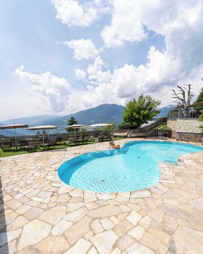 Piscina con baldosas de piedra, mobiliario exterior y vistas a montañas y un lago.