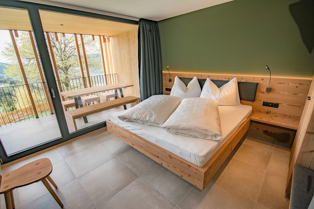 A bedroom with a wooden bed, a wooden nightstand, a bench, and a view of the outdoors.