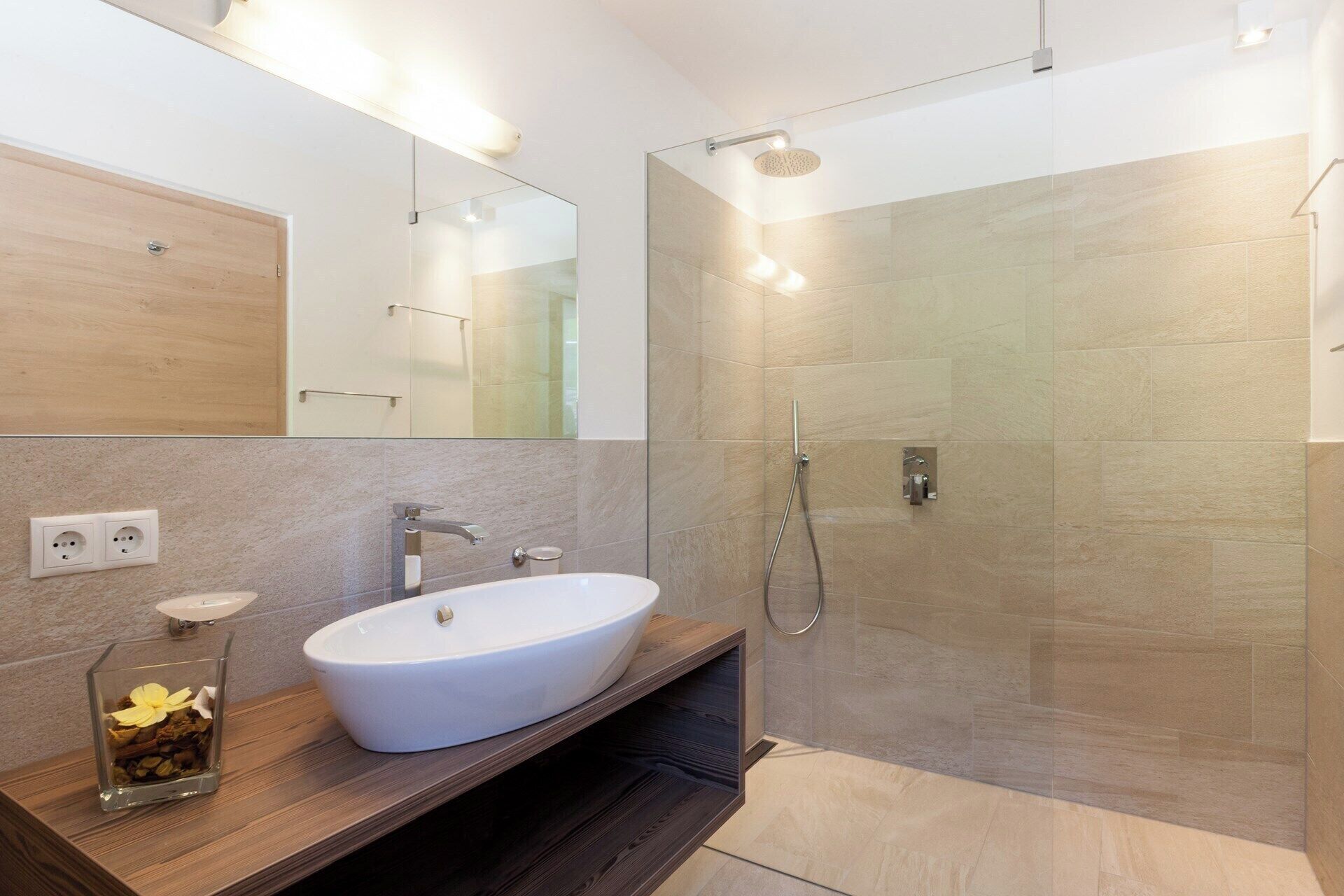 A modern bathroom with a large white sink, a glass vase with flowers, a mirror, and a glass-enclosed shower area.