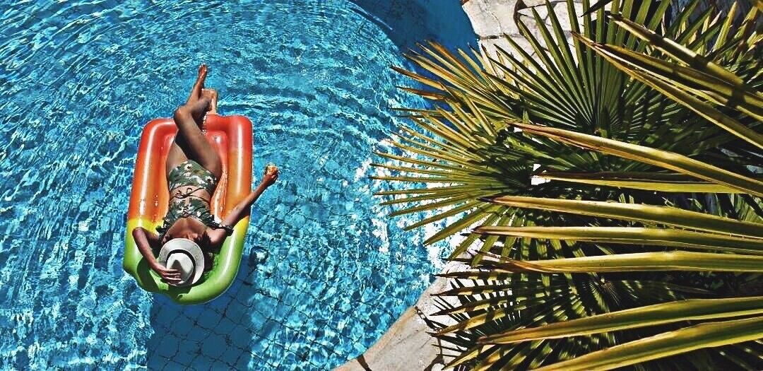 Una persona recostada sobre un flotador inflable de colores en una piscina azul cristalina, rodeada de hojas de palmera.