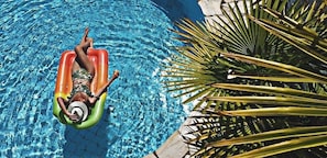 Una persona estirada sobre un salvavides inflable i colorit en una piscina cristal·lina i blava, envoltada de fulles de palmera.
