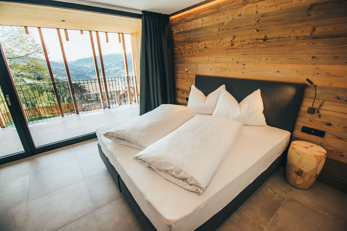 A modern bedroom with a large bed, wooden panel walls, and a balcony with a view of the mountains.
