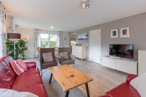 A modern living room with a red leather sofa, a wooden coffee table, and a flat-screen TV. There is a dining area with a white cabinet and a kitchen in the background.