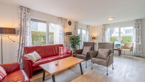 A spacious living room with a red leather sofa, a wooden coffee table, and a brown leather armchair. There are two floor lamps, a potted plant, and a view of the outside through large windows. - Charming Villa Near the sea and Forest (De Koog)
