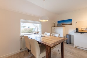 A dining area with a wooden table and white chairs, a window with a view, a radiator, a trash bin, and a kitchen area with cabinets and a countertop.