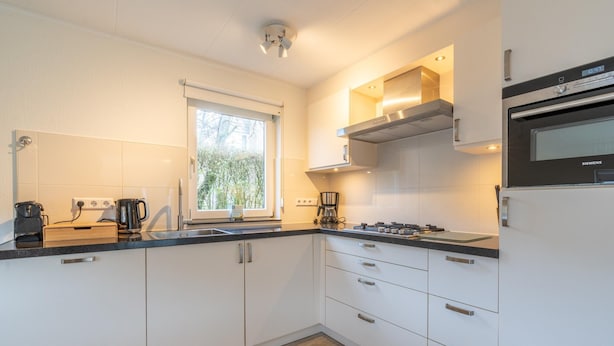 A modern kitchen with white cabinets, a built-in oven, and a window providing natural light. - Charming Villa Near the sea and Forest (De Koog)