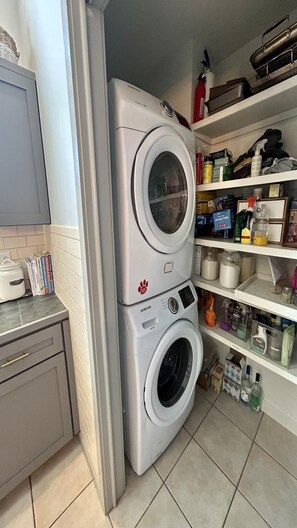 A laundry area with stacked washing machines, a shelf with cleaning supplies, and a tiled floor.