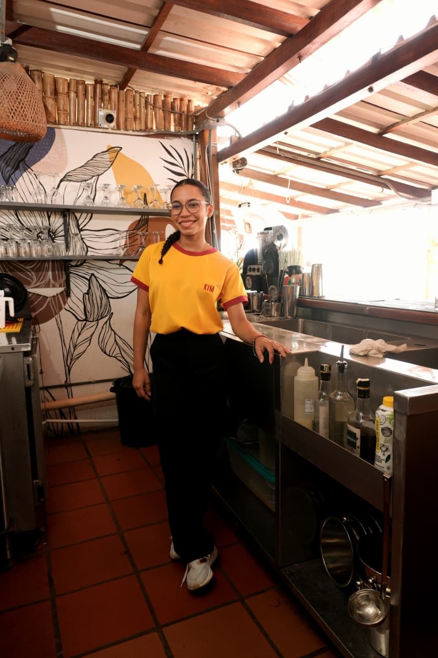 Un coin cuisine avec une personne debout près du comptoir, des étagères remplies de verres et une fresque murale.