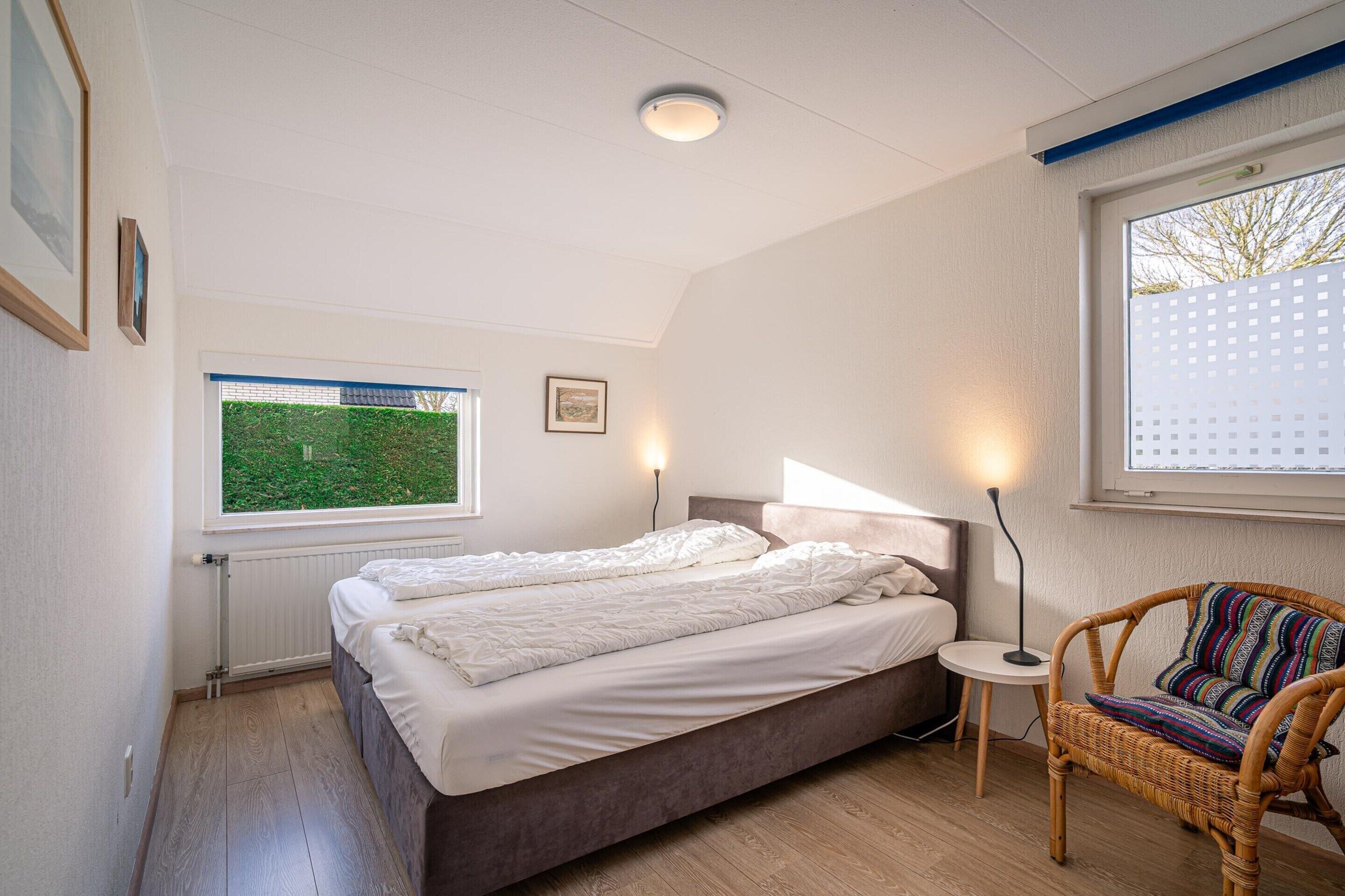 Une chambre à coucher avec un lit, une fenêtre donnant sur un paysage verdoyant, une chaise en osier et une table de chevet avec une lampe.