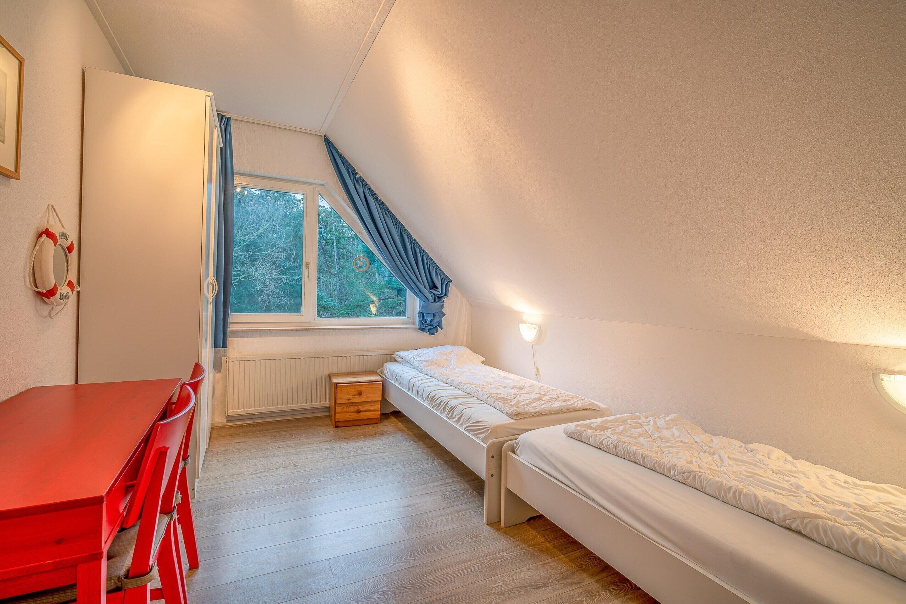 A small, well-lit room with a single bed, a red table, and a window with curtains.