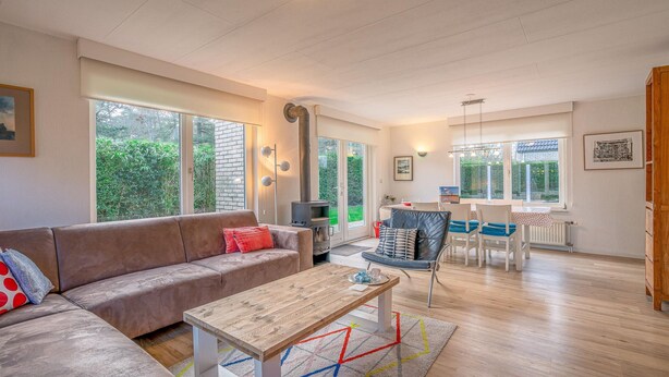 A spacious living area with a sofa, a coffee table, and a dining table with chairs. There are large windows providing natural light and views of greenery outside. - Charming Villa Near the sea and Forest (De Koog)