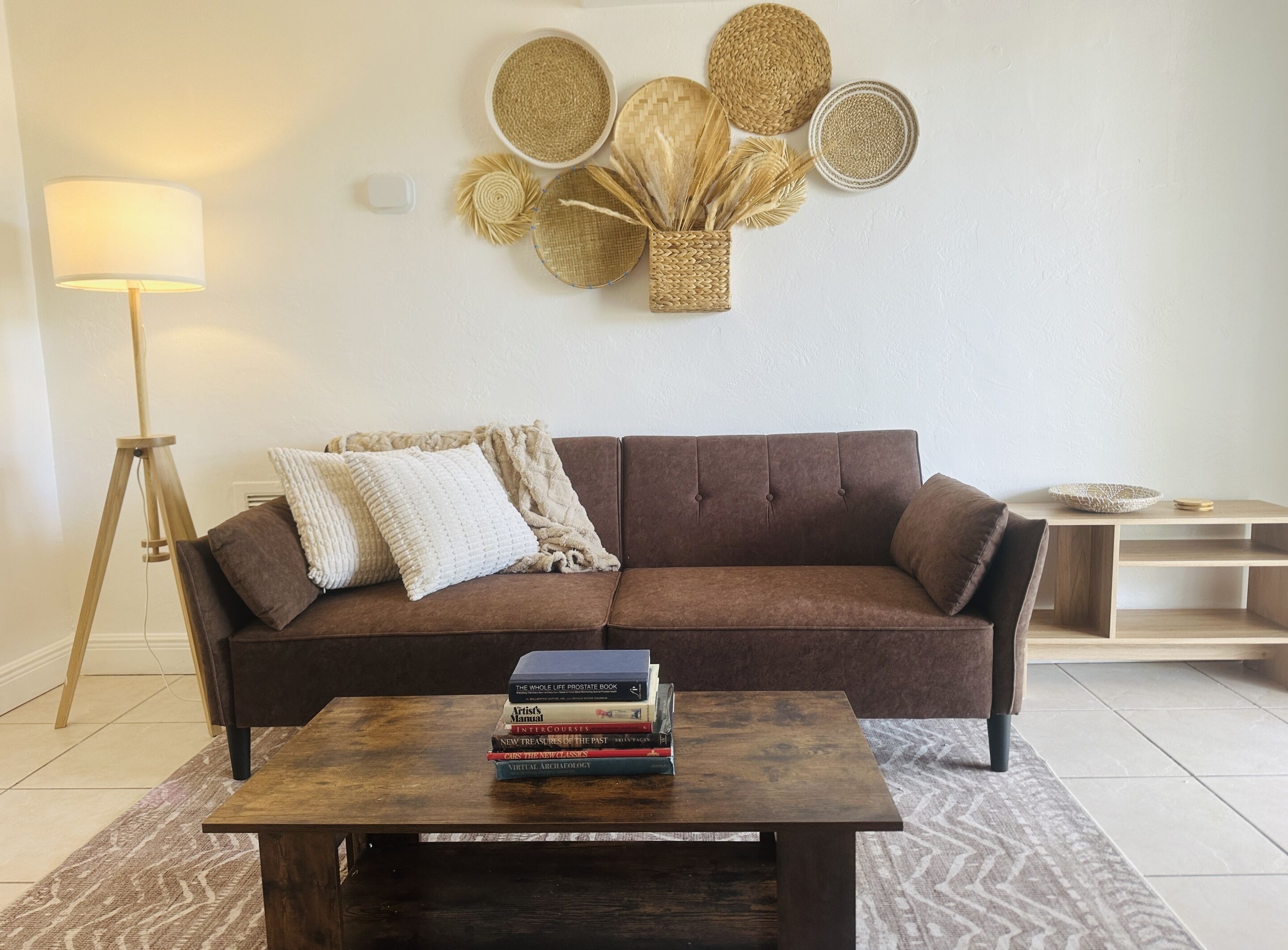 Una sala de estar con un sofá marrón, una mesa de centro de madera con libros, una lámpara de pie y un adorno de pared tejido.