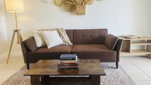 A living room with a brown sofa, a wooden coffee table with books, a floor lamp, and woven wall art. - Wilton Manors Guesthouse #1 (Wilton Manors)