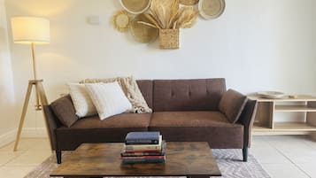 Una sala de estar con un sofá marrón, una mesa de centro de madera con libros, una lámpara de pie y un adorno de pared tejido.