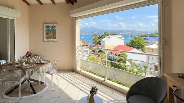 Un balcon avec vue sur la mer et les bâtiments, une table à manger dressée avec des assiettes et des tasses, et une chaise.