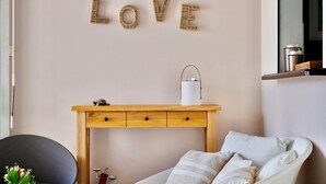 Une pièce dans laquelle on peut voir, accroché au mur, le mot « AMOUR » écrit en lettres dorées, une table d’appoint en bois sur laquelle est posé un vase blanc, une chaise grise et un canapé blanc agrémenté de coussins à rayures.