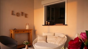 Une pièce chaleureuse avec un canapé rond blanc, une petite table sur laquelle est posé un vase de fleurs, une table d’appoint en bois et une étagère murale agrémentée d’un miroir.