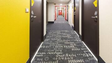 Un pasillo de hotel con una alfombra que presenta texto y símbolos, puertas con letreros amarillos y una alfombra roja al final.