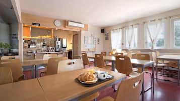 A dining area with multiple tables and chairs, a kitchen in the background, and a clock on the wall.
