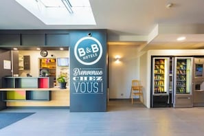 A hotel reception area with a B&B logo, a welcome sign, a reception desk, vending machines, and a seating area. - B&B Hotel Valence Sud (Valence)