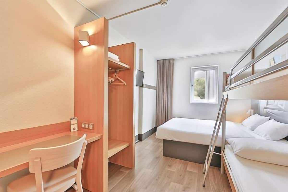 A hotel room with a bunk bed, a desk with a chair, a wardrobe, and a window with curtains.