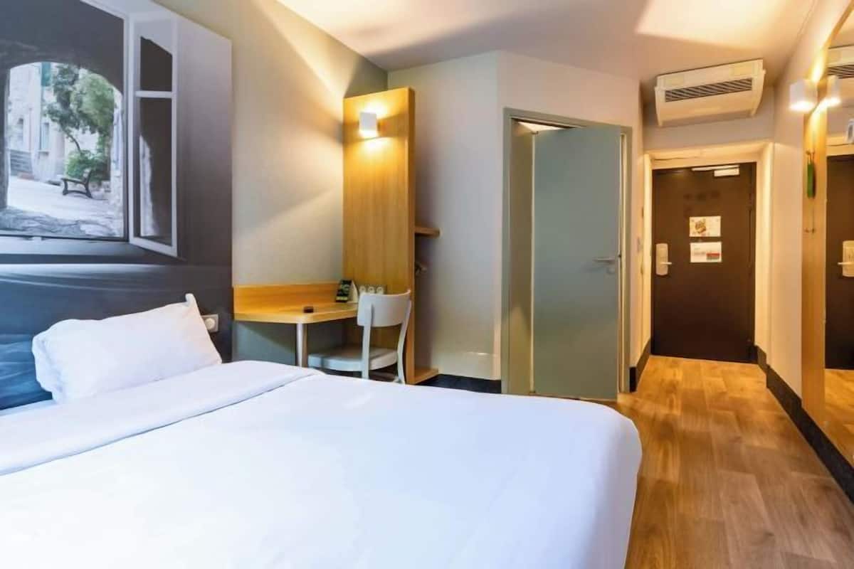 A hotel room with a bed, a desk, a chair, a door, and a hallway.