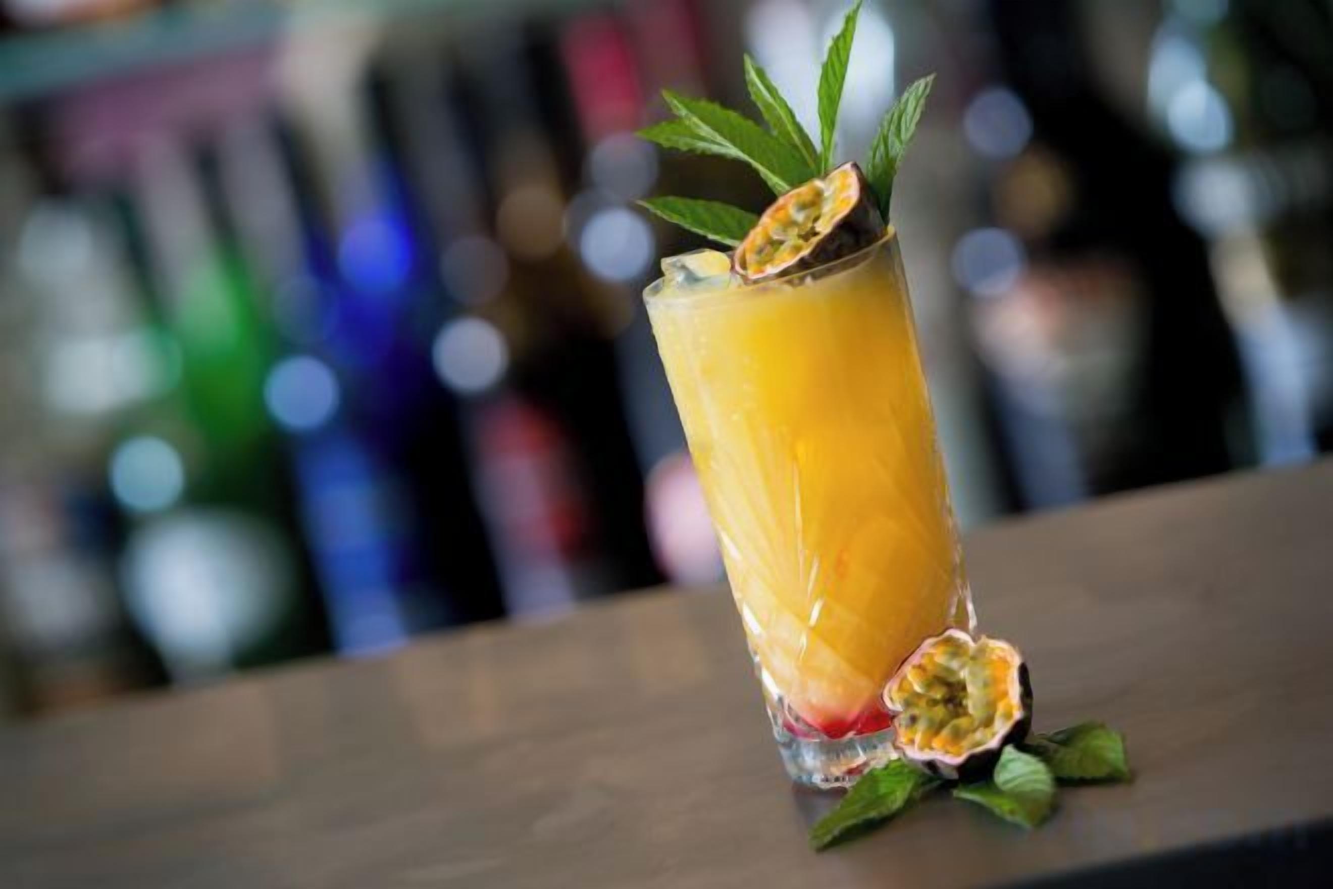 Een hoog glas met een geel drankje, versierd met een blaadje en een stuk fruit, op een barkruk.