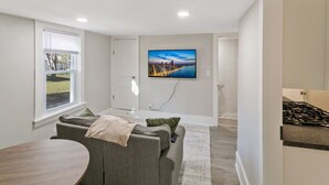 Una sala moderna con un televisor de pantalla plana montado en la pared, un sofá gris con una manta, una mesa de comedor de madera y una ventana con persianas.