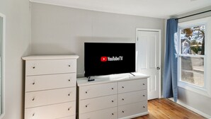 A room with a white dresser, a flat-screen TV displaying YouTubeTV, a door, and a window with curtains. - Cozy Spot in Heart of Libertyville Near Naval Base (Libertyville)
