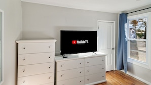 Quarto com cômoda branca, televisão de tela plana exibindo YouTubeTV, porta e janela com cortinas.