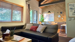 A cozy living room with a dark leather sofa, wooden beams on the ceiling, and a large window with blinds. - NEW & FUN! Hot Tub, EV Outlet in Garage (Mammoth Lakes)