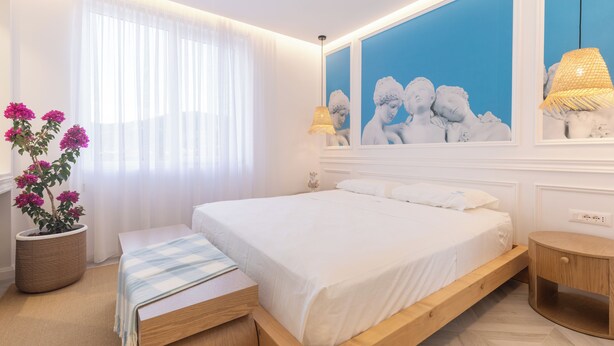 Une chambre à coucher avec un lit, une table de chevet, une lampe et une grande fenêtre ornée de rideaux légers.
