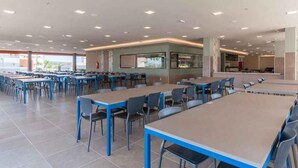A spacious cafeteria with multiple tables and chairs, a counter area, and a large window providing a view of the outdoors.