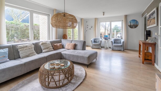 A spacious living room with a grey sectional sofa, a wicker coffee table, and a large window offering a view of the outdoors. - Charming Villa Near the sea and Forest (De Koog)