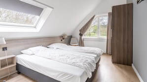 A bedroom with two beds, a skylight, a window with curtains, a bedside lamp, and a wooden wardrobe. - Charming Villa Near the sea and Forest (De Koog)