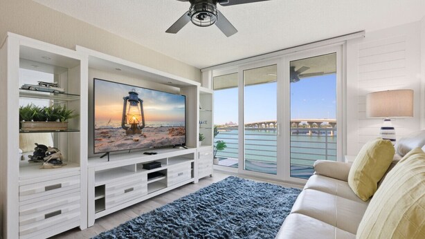 Una sala de estar con un televisor de pantalla plana grande, un ventilador de techo, un sofá y vistas a una masa de agua a través de amplias ventanas.