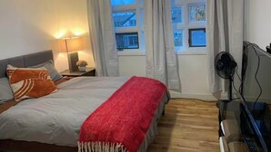 Et soveværelse med seng, et sengebord med lampe, et fjernsyn og et vindue med gardiner.