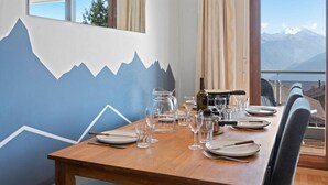 A dining table set with plates, glasses, and cutlery, with a mountain mural on the wall and a view of mountains through the window. - Crans-Montana, 5 pers : balcon vue & parking privé (Crans-Montana)