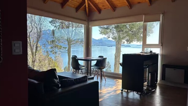 A cozy room with a wooden ceiling, a dining area with chairs and a table, and a large window overlooking a lake and mountains. - Upstairs apartment - Seeblick Bariloche (San Carlos de Bariloche)