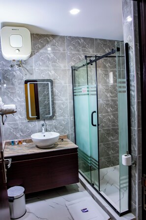 A modern bathroom with a marble-tiled wall, a glass-enclosed shower, a white sink, and a mirror. - Villa Hilton Hotels & Apartments (Uyo)