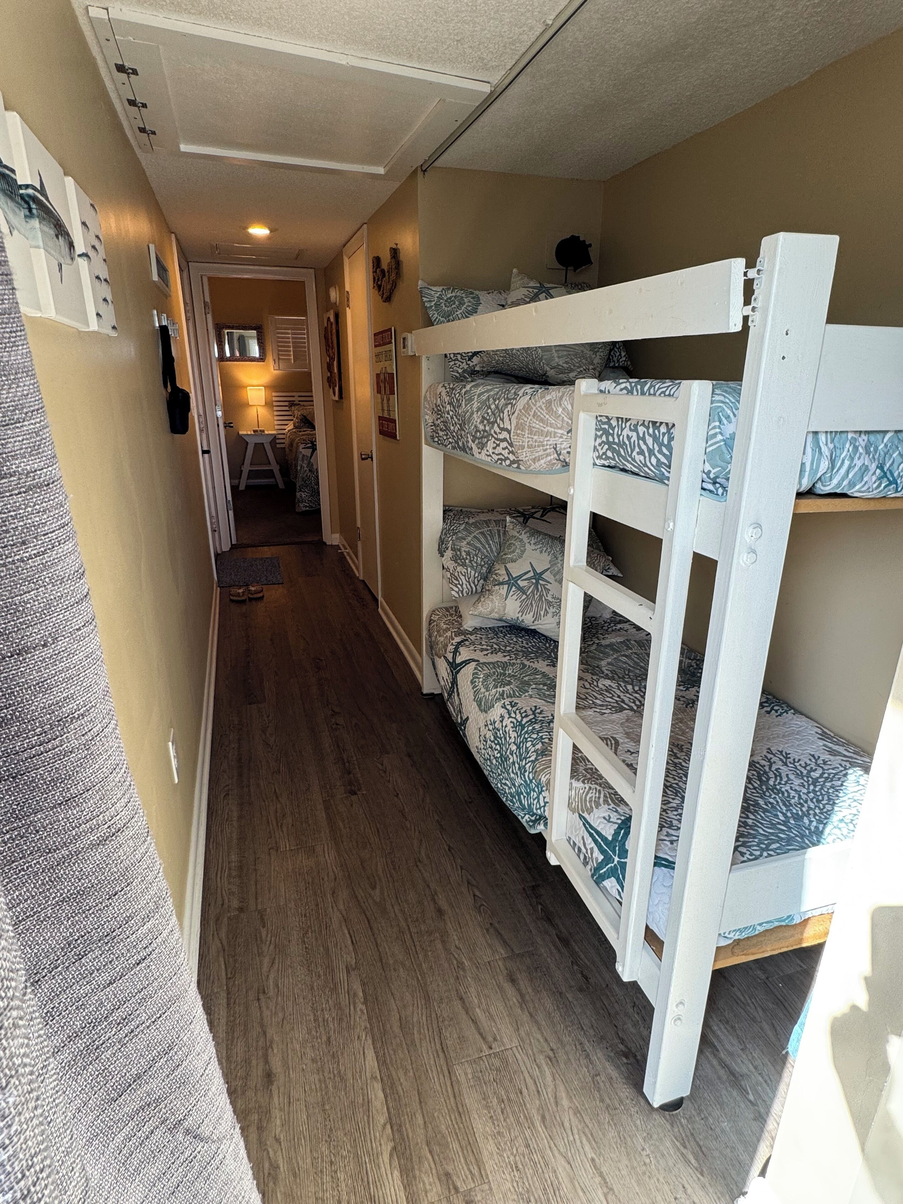 A narrow hallway with bunk beds on the right, wooden flooring, and a door at the end leading to a room with a bed and a lamp.